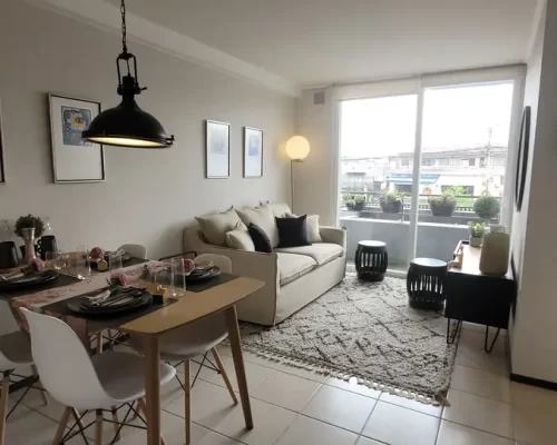 Vista del área de living comedor amoblada con un sofá beige, alfombra de diseño geométrico y acceso directo a un balcón iluminado.