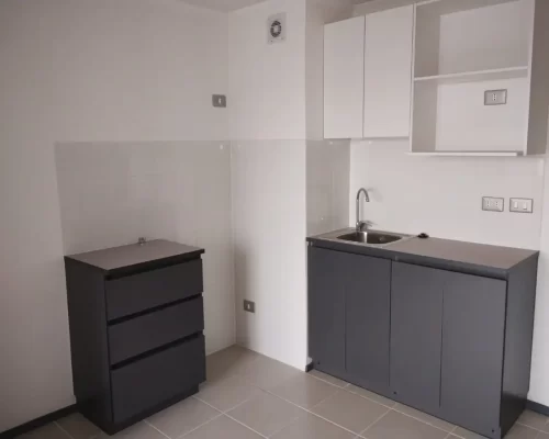 Cocina equipada con muebles inferiores grises, encimera clara, lavamanos de acero inoxidable y repisas superiores en blanco.
