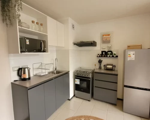 Cocina equipada con muebles inferiores grises, encimera de melamina, cocina a gas, microondas integrado en estantería superior y un refrigerador de acero inoxidable.