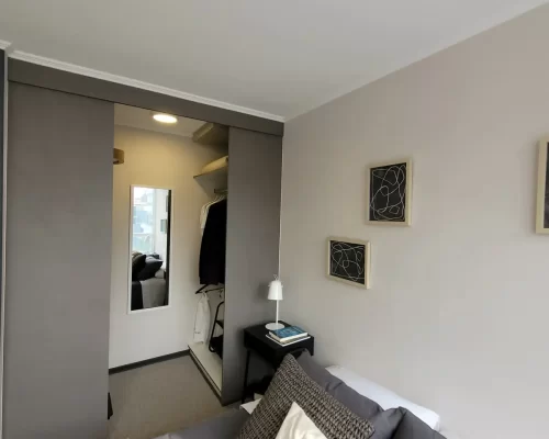 Vista del dormitorio que muestra un clóset empotrado con puertas correderas en color gris oscuro, revelando un espacio interior para colgar ropa y un espejo de pared.