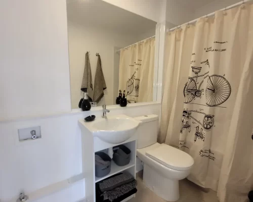 Baño moderno con lavamanos sobre mueble blanco con repisas de organización, espejo de muro a muro y tina con cortina decorativa con diseño de bicicleta vintage.