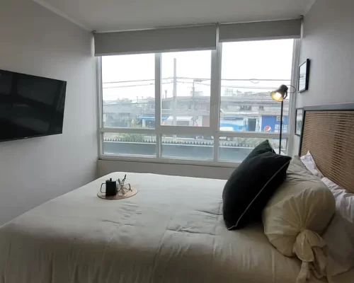 Dormitorio principal con cama matrimonial, televisor empotrado en la pared y un gran ventanal de termopanel con cortinas roller.